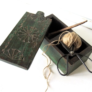 Puede incluir: Una caja de madera verde oscuro con un dise&ntilde;o floral tallado en la tapa. En el interior, un ovillo de hilo, unas tijeras negras y un palito de madera son visibles. La caja est&aacute; abierta, revelando su contenido.