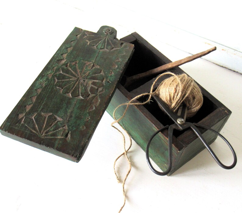 Puede incluir: Una caja de madera verde oscuro con un dise&ntilde;o floral tallado en la tapa. En el interior, un ovillo de hilo, unas tijeras negras y un palito de madera son visibles. La caja est&aacute; abierta, revelando su contenido.