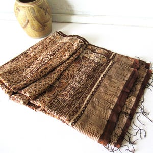 May include: A rectangular textile featuring a brown and tan batik pattern with floral motifs and geometric borders. The fabric has a striped section on one end and dark brown fringe along the edges, displaying a complex, layered design.