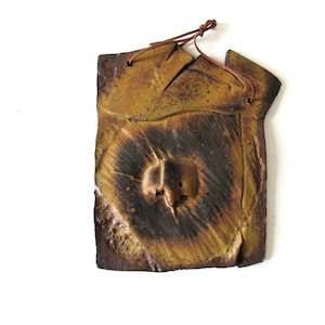 May include: A rectangular ceramic wall plaque with a textured, mustard-yellow and dark brown glaze. The surface features a central raised protrusion and radiating lines. A thin brown leather cord is threaded through two holes at the top for hanging.