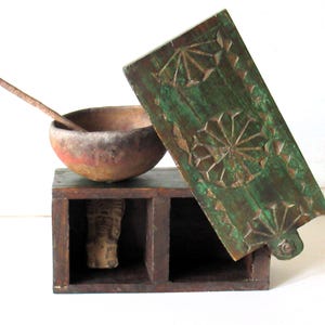 Puede incluir: Una caja de madera antigua con tapa con bisagras, un cuenco peque&ntilde;o y una figura de madera tallada. La caja es de color marr&oacute;n oscuro con una tapa verde con dise&ntilde;os geom&eacute;tricos tallados. El cuenco es de color marr&oacute;n rojizo.