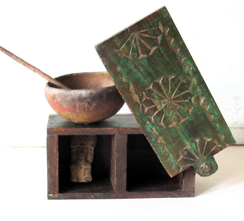 Puede incluir: Una caja de madera antigua con tapa con bisagras, un cuenco peque&ntilde;o y una figura de madera tallada. La caja es de color marr&oacute;n oscuro con una tapa verde con dise&ntilde;os geom&eacute;tricos tallados. El cuenco es de color marr&oacute;n rojizo.
