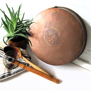 May include: A large, antique copper pan with a dark patina, resting on a white surface. A wooden spoon and a slotted utensil are next to a black and white striped cloth. A potted aloe vera plant adds a touch of green.