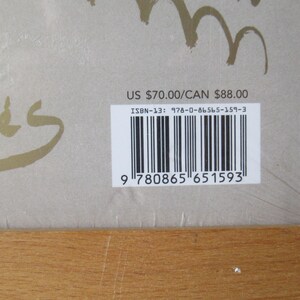 May include: A book cover with gold script and a barcode. The cover is a light tan color with the word "m&eacute;s" in gold script. A barcode and ISBN-13 number are visible on a white sticker.