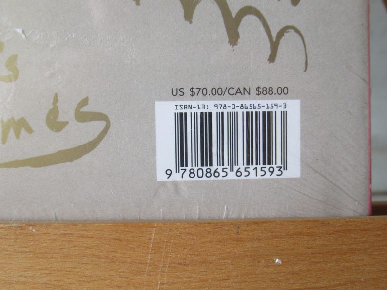 May include: A book cover with gold script and a barcode. The cover is a light tan color with the word "m&eacute;s" in gold script. A barcode and ISBN-13 number are visible on a white sticker.