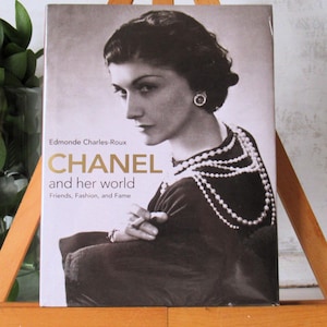 May include: A book titled "CHANEL and her world" featuring a black and white portrait of Coco Chanel. The cover includes the author's name, Edmonde Charles-Roux, and the subtitle "Friends, Fashion, and Fame."
