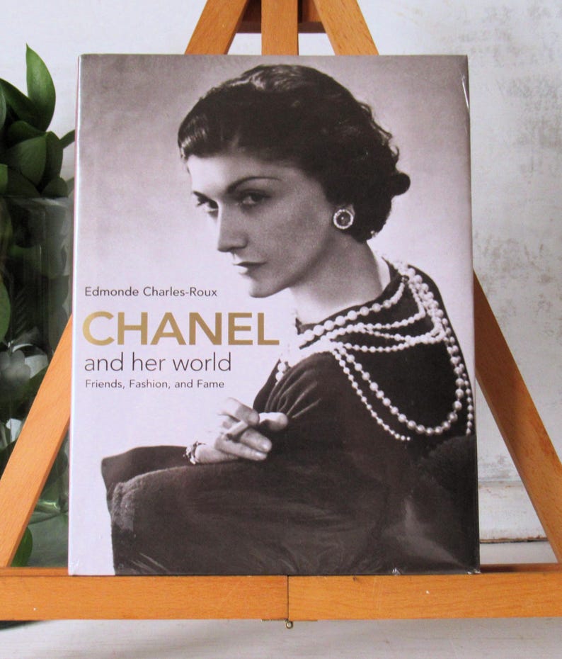 May include: A book titled "CHANEL and her world" featuring a black and white portrait of Coco Chanel. The cover includes the author's name, Edmonde Charles-Roux, and the subtitle "Friends, Fashion, and Fame."
