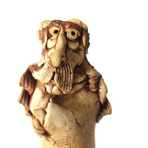 May include: A hand-sculpted ceramic bust featuring a textured, light brown clay finish. The figure displays a prominent nose, a detailed moustache, and deep-set eyes, with layered, wavy hair framing the face and a draped garment over the shoulders.