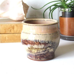 May include: A small, round ceramic pot featuring a variegated glaze with horizontal bands of cream, tan, and deep brown. The vessel has a smooth, slightly flared rim and a tapered base, showcasing a reactive drip pattern that blends earthy tones.