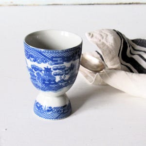 May include: A blue and white ceramic egg cup with a detailed Willow pattern design. The cup has a pedestal base and a wide rim. A silver spoon rests on a white and black striped napkin.