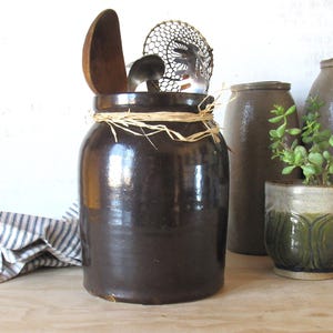 May include: A dark brown cylindrical stoneware crock featuring a smooth, glazed finish and a natural raffia tie wrapped around the neck. The crock contains a wooden spoon, a metal ladle, and a metal pasta server, with a striped cloth nearby.