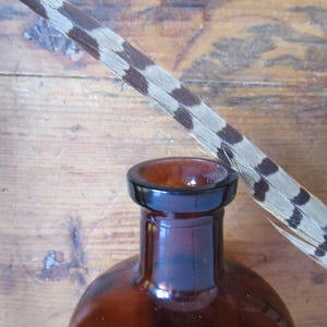 May include: A brown glass bottle with a black rim and a feather with brown and white markings. The feather is angled across the top of the bottle, set against a wooden background.