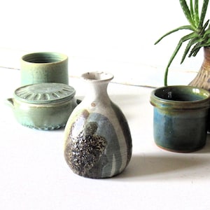 May include: A small, bulbous ceramic vase with a narrow neck, featuring a mottled grey, white, and dark brown textured glaze. It is surrounded by other ceramic vessels, including a lidded green container and a blue-green cylindrical pot.