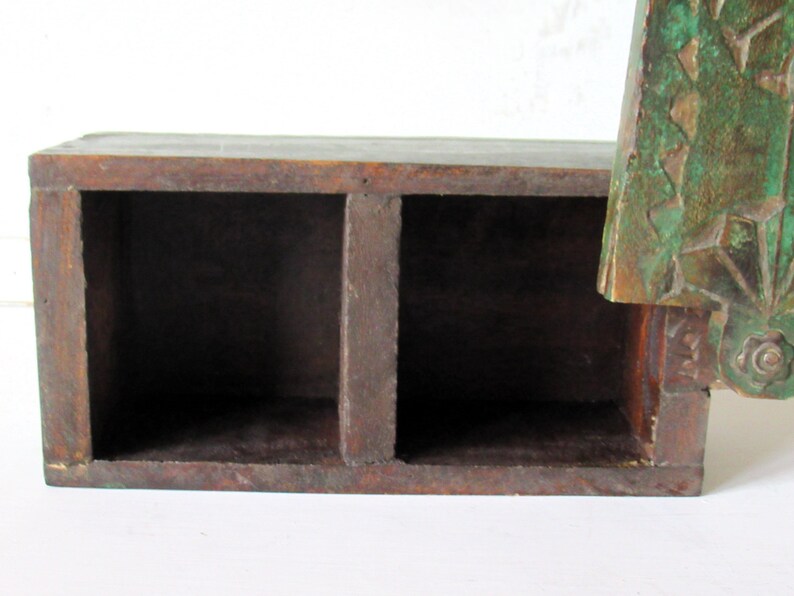 Puede incluir: Una caja de madera marr&oacute;n oscuro con dos compartimentos. La caja tiene forma rectangular y parece hecha a mano. Un elemento decorativo verde y bronceado est&aacute; unido al lateral, con dise&ntilde;os florales y geom&eacute;tricos.