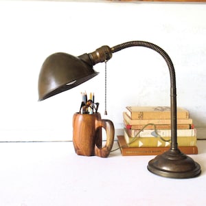 May include: An antique brass desk lamp with a flexible neck and a dark green shade. A wooden mug filled with pencils sits beneath the lamp. A stack of vintage books is to the right.