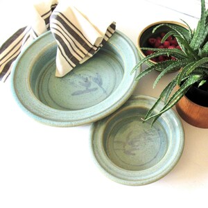 Puede incluir: Dos cuencos redondos de cerámica azul claro con un acabado moteado y un sutil diseño floral azul en el centro. Una servilleta de tela blanca con rayas de color marrón oscuro cubre el cuenco más grande y hay una planta de aloe verde cerca.