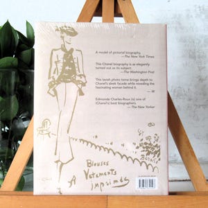 May include: A book with a light pink cover featuring a sketch of a person in a hat and suit. Text includes quotes from The New York Times, The Washington Post, and The New Yorker, along with the words "Blouses Vetements imprim&eacute;s."