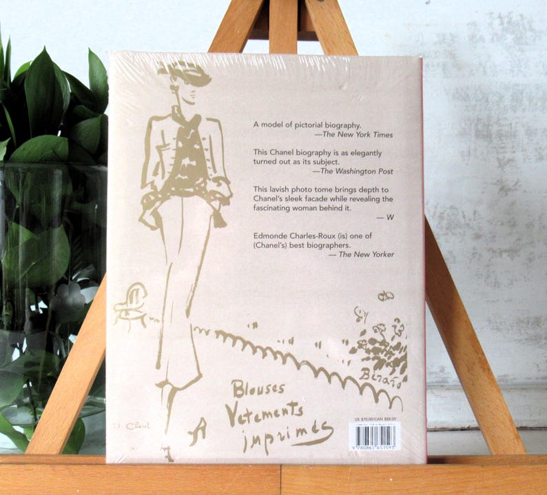 May include: A book with a light pink cover featuring a sketch of a person in a hat and suit. Text includes quotes from The New York Times, The Washington Post, and The New Yorker, along with the words "Blouses Vetements imprim&eacute;s."