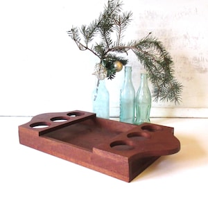 Puede incluir: Una bandeja de madera para servir con un acabado marrón rojizo intenso. La bandeja presenta varios recortes circulares para sostener vasos o botellas. Ramas decorativas de hoja perenne y botellas de vidrio vintage en el fondo.