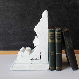 May include: A white painted wooden corbel bookend with a curved, ornate profile positioned next to three dark green and black hardcover books. The books are titled Longfellow Works Outre-Mer, Longfellow Works Hyperion, and The Essays of Ralph Waldo Emerson.
