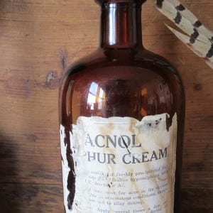 May include: A vintage brown glass bottle with a partially peeled label. The label reads "ACNOL PHUR CREAM" in bold letters. A feather with brown and black markings rests near the bottle, suggesting a rustic or antique theme.
