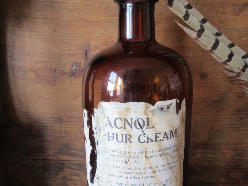 May include: A vintage brown glass bottle with a partially peeled label. The label reads "ACNOL PHUR CREAM" in bold letters. A feather with brown and black markings rests near the bottle, suggesting a rustic or antique theme.