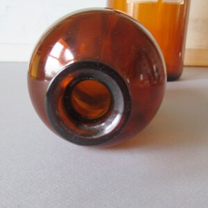 May include: Close-up of a brown glass bottle with a round body and a dark brown neck. The bottle is empty and has a smooth, reflective surface. Another bottle is visible in the background. The bottles are likely for storing liquids.