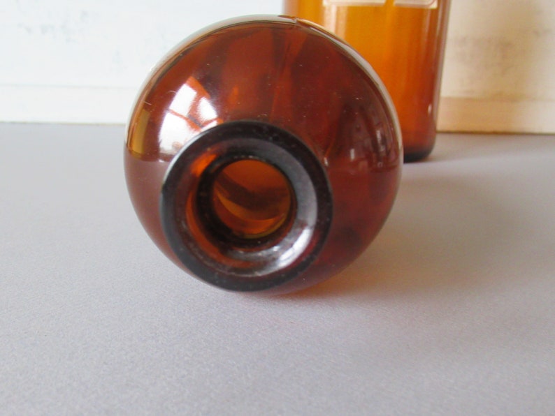 May include: Close-up of a brown glass bottle with a round body and a dark brown neck. The bottle is empty and has a smooth, reflective surface. Another bottle is visible in the background. The bottles are likely for storing liquids.