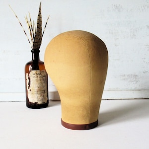 May include: A tan-colored wig head form with a brown base. A vintage brown glass bottle with a label and feathers is in the background. The wig form is made of a soft fabric and is designed to hold wigs.