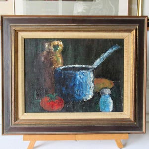 May include: An oil painting in a dark wood frame depicting a still life scene. The composition features a brown jug, a bright blue saucepan with a long handle, a red tomato, a small blue shaker, and an orange object, all set against a dark background.