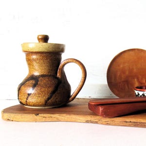 May include: A brown ceramic pitcher with a lid and a curved handle, featuring a speckled texture and dark brown vertical brushstroke accents. The pitcher sits alongside a round wooden plate and a long, smooth wooden object on a wooden board.