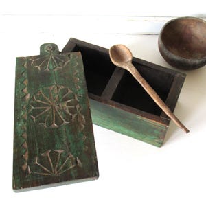 Puede incluir: Una caja de especias de madera verde r&uacute;stica con un dise&ntilde;o floral tallado en la tapa. La caja est&aacute; abierta, revelando dos compartimentos, e incluye una cuchara de madera. Tambi&eacute;n hay un cuenco de madera peque&ntilde;o.