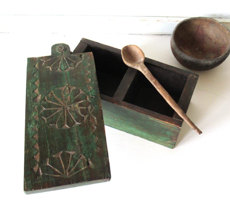 Puede incluir: Una caja de especias de madera verde r&uacute;stica con un dise&ntilde;o floral tallado en la tapa. La caja est&aacute; abierta, revelando dos compartimentos, e incluye una cuchara de madera. Tambi&eacute;n hay un cuenco de madera peque&ntilde;o.