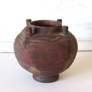 May include: A handcrafted, brown ceramic vase with a textured, wave-like pattern. The vase has a rounded body, a small pedestal base, and four small, cylindrical handles near the rim. The colors include shades of brown, red, and green.