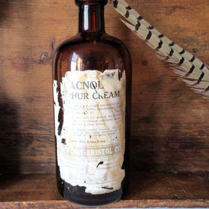 May include: An antique brown glass bottle with a peeling label reading "ACNOL PHUR CREAM." The label has faded text and is partially torn. A feather with brown and cream markings rests against the bottle. The bottle is set against a wooden background.