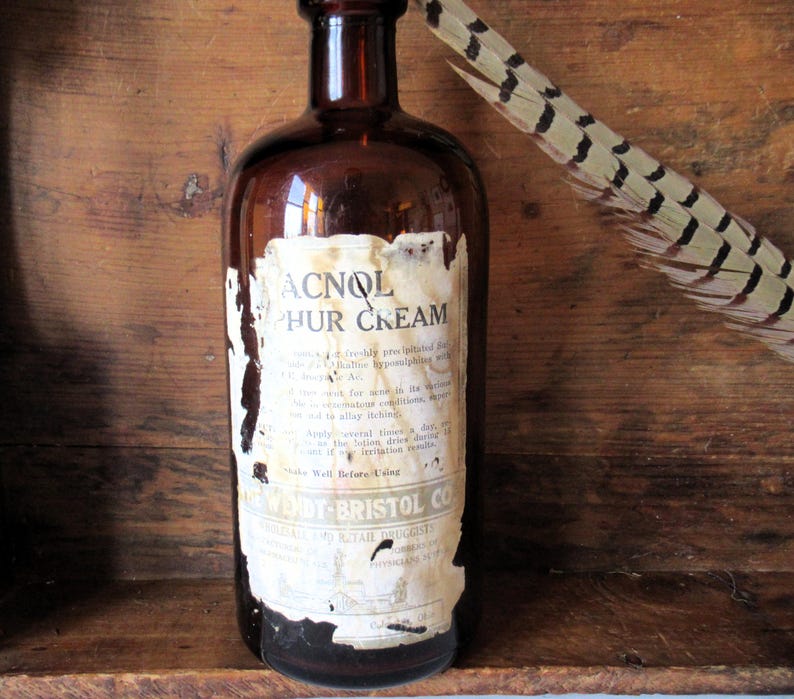 May include: An antique brown glass bottle with a peeling label reading "ACNOL PHUR CREAM." The label has faded text and is partially torn. A feather with brown and cream markings rests against the bottle. The bottle is set against a wooden background.
