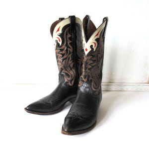 Puede incluir: Un par de botas vaqueras de cuero negro con punta afilada y caña alta. La caña está decorada con costuras intrincadas en tonos marrón, crema y rojo, con un distintivo aplique de cuero color crema en la parte superior del borde.