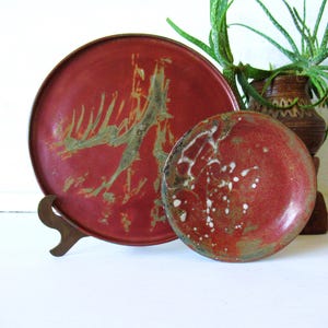 May include: Two round ceramic plates with a deep red glaze and textured, earthy green and white markings. The larger plate is supported by a metal stand, while the smaller plate sits in front, both displaying a mottled, organic finish.