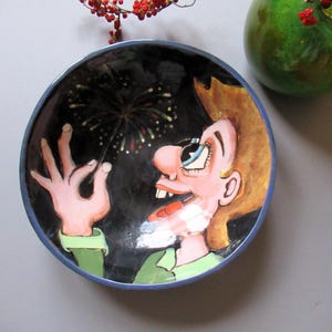 May include: A hand-painted ceramic bowl with a whimsical illustration of a person watching fireworks. The bowl has a black interior with a blue rim and a green shirt. The person has brown hair and a surprised expression.