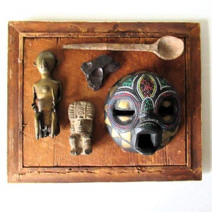 May include: A rectangular wooden frame containing a collection of objects: a metallic standing figure, a small stone carving, a dark rock fragment, a wooden spoon, and a round black mask decorated with colorful beads and gold triangular accents.