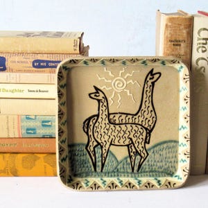 May include: A square ceramic plate with a tan base color featuring a black line drawing of two llamas standing in a field under a stylized sun. The border of the plate is decorated with a repeating green and black geometric pattern.