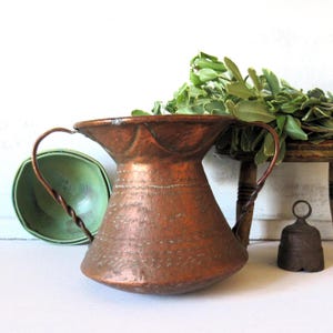 May include: A copper pitcher with a wide mouth and two twisted handles. The pitcher has a textured surface with etched details. A small green bowl and a small bell are next to the pitcher. Green leaves are on a wooden stool in the background.