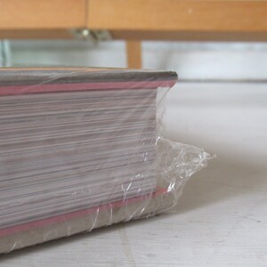May include: A stack of paper wrapped in clear plastic. The edges of the paper are visible, with a red stripe along the side. The paper is stacked on a white surface, with a wooden structure in the background.