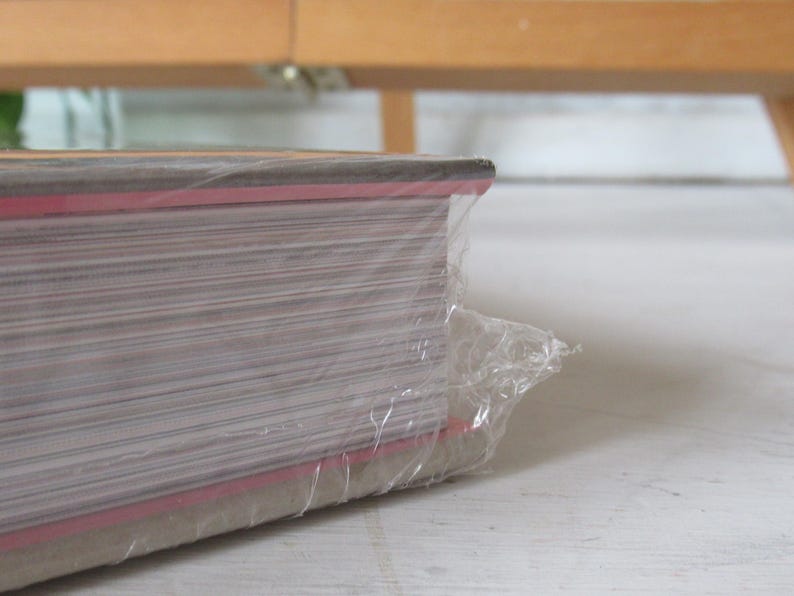 May include: A stack of paper wrapped in clear plastic. The edges of the paper are visible, with a red stripe along the side. The paper is stacked on a white surface, with a wooden structure in the background.