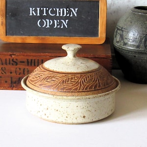 May include: A ceramic lidded serving dish with a leaf pattern on the lid. The dish is off-white with brown accents and speckles. A wooden framed chalkboard with the words "KITCHEN OPEN" is in the background.