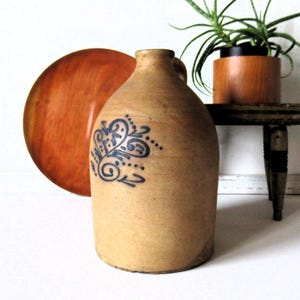 May include: A tan stoneware jug with a narrow neck and a single handle, featuring a dark blue decorative floral pattern painted on the front. The jug has a cylindrical body with a flat base and a smooth, matte, earthy-toned surface texture.