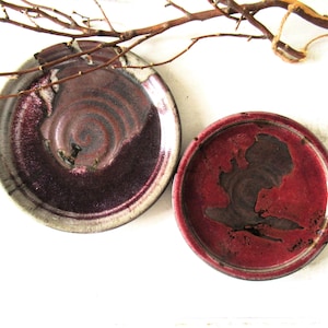 May include: Two circular ceramic plates featuring a glossy glaze in shades of deep burgundy, plum, and dark red. The larger plate has a light grey rim with a spiral pattern in the center, while the smaller plate displays a solid dark red interior.