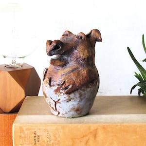 May include: A handcrafted ceramic pig figurine with a brown and tan finish. The pig has a detailed snout, ears, and paws, and is sitting on a book. The figurine is a unique decorative item.