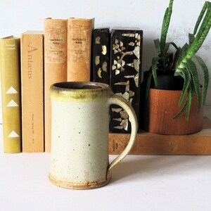 May include: A tall, cylindrical ceramic mug with a speckled off-white glaze and a darker, yellowish-brown rim. The mug features a sturdy, curved handle on the right side and a slightly flared base, showcasing a handcrafted, textured appearance.