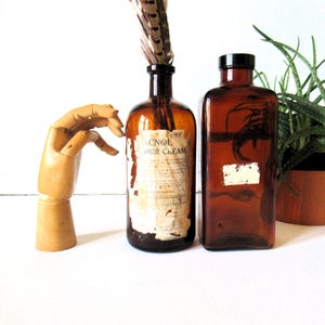 May include: Two antique amber glass bottles, one with a label reading "ACNOL HUR CREAM" and a feather, the other with a blank label. A wooden articulated hand is on the left. A potted plant is on the right.